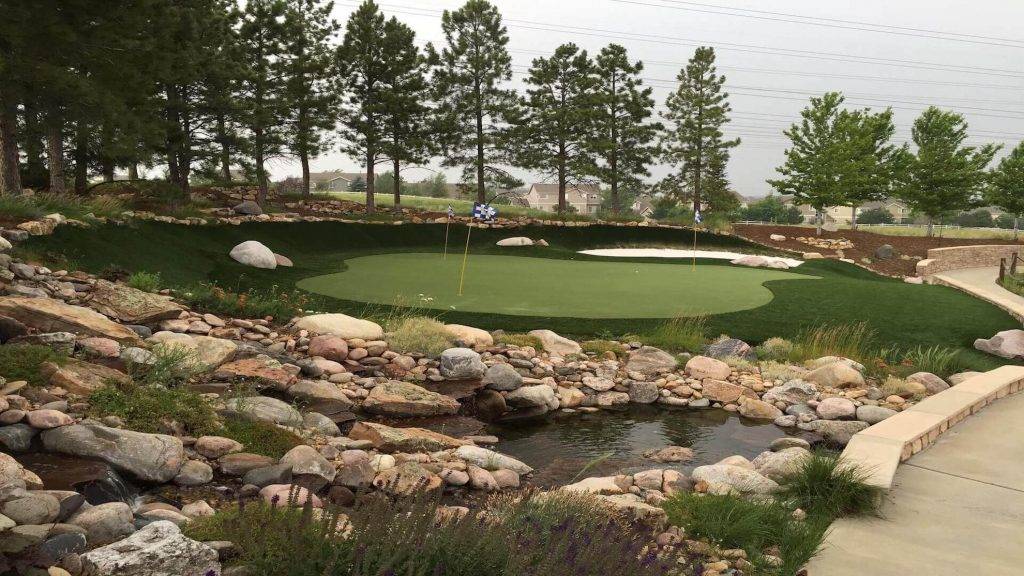 Putting green with two sand traps and pond close by. Cloudy skies and pine trees in the background.